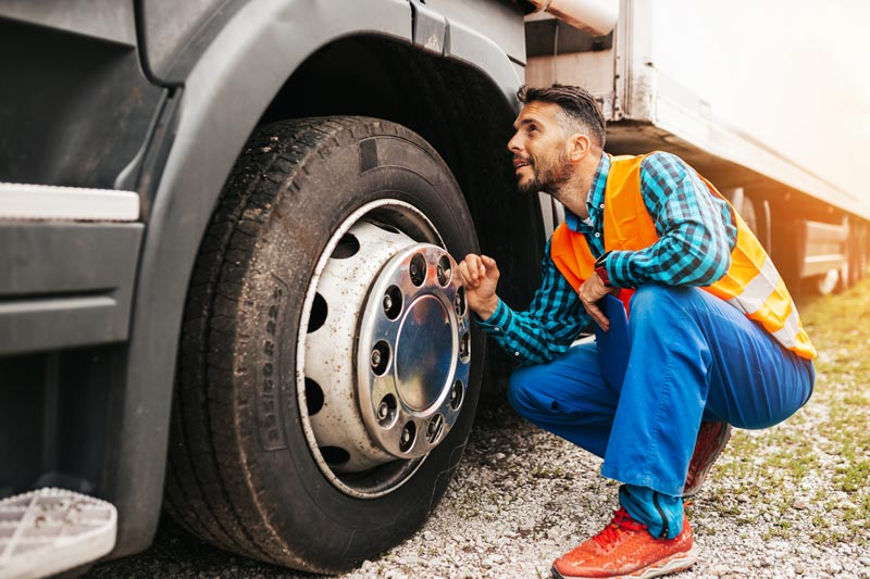 A man inspects a truck tire, crouching on gravel