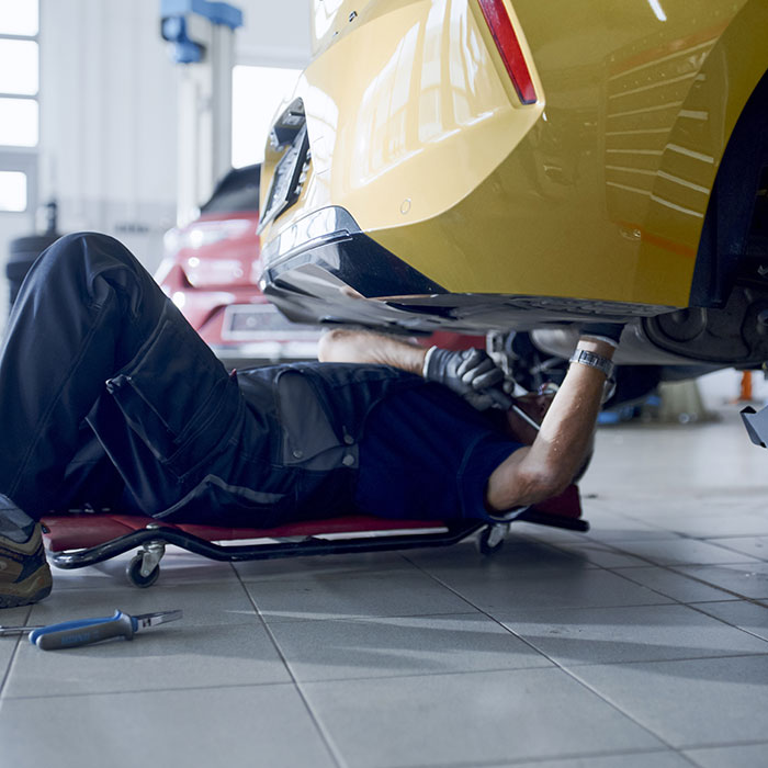 A mechanic in blue overalls lies on a creeper under a yellow car