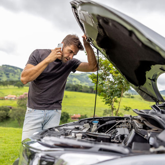 A man stands by an open car hood on a rural road, speaking on a phone